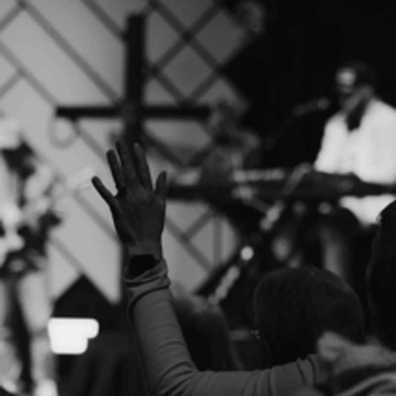A black and white photo of a person from behind with their hand raised in the air, facing a stage with a band and a cross during a church service.
